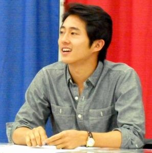 Steven Yeun sitting at a table in front of a curtain