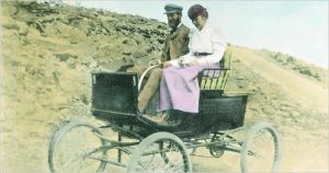 A man and woman driving a horseless carriage